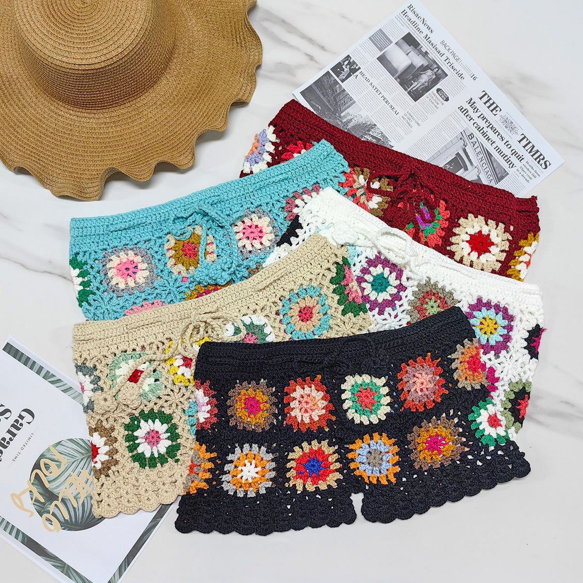 A collection of women's beach shorts in various colors, featuring a crocheted floral pattern, displayed on a surface with a straw hat and a newspaper for added context.