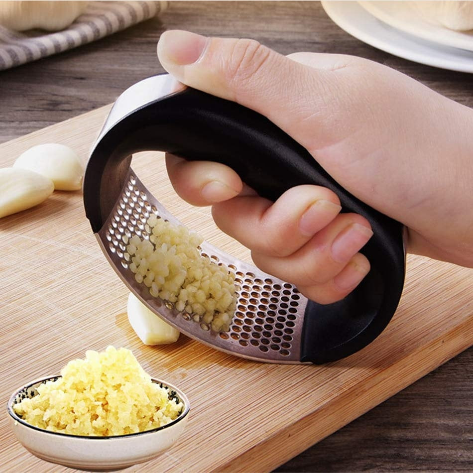 A stainless steel garlic press being used to mash garlic on a wooden cutting board with a non-slip black handle.