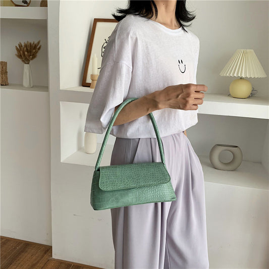 A person holding a green handbag with a solid pattern, in a room with a white wall and a shelf with decorative items.