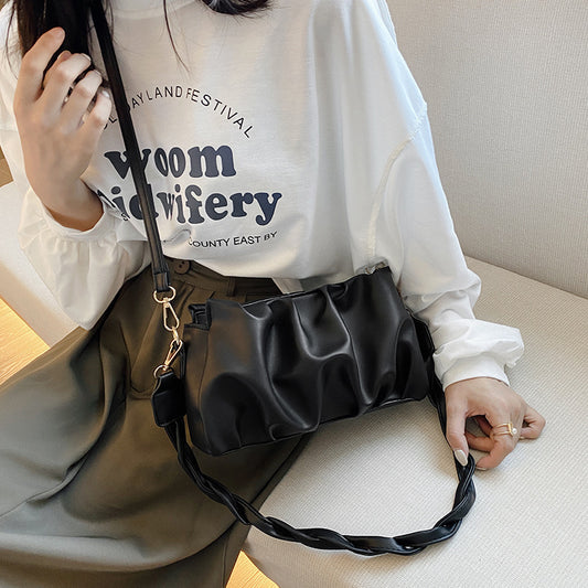 A woman holding a black pleated shoulder bag with a single strap over her shoulder, wearing a white long-sleeve top with text and beige pants.