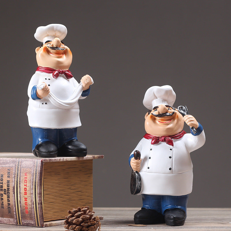 Two resin statues of cartoonish chefs in white uniforms, one standing and the other sitting, both with a chef's hat and apron, posed with a towel and a spoon.