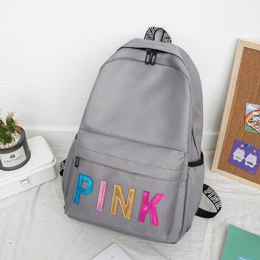 A gray women's backpack with 'PINK' written in colorful sequins on the front. The backpack features a zippered opening and it is placed on a bed next to a plant and some stationery items.