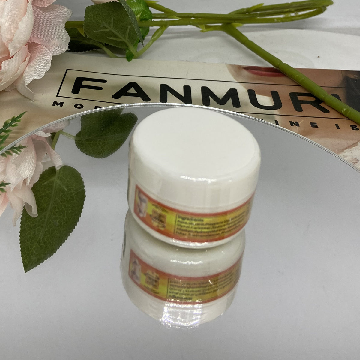 White jar with labels on a reflective surface, with flowers and leaves in the background.