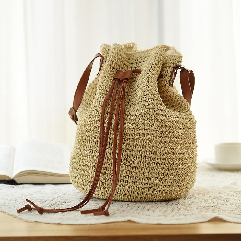 A beige ethnic style drawstring woven straw bag with a polyester texture, displayed on a table with a soft background.