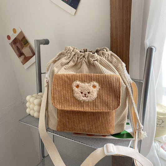 A khaki corduroy drawstring bucket bag with a bear face design on the front, displayed on a metal rack.