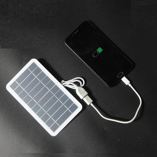 A portable solar panel charger connected to a black smartphone via a USB cable, with the solar panel laid flat on a dark surface.