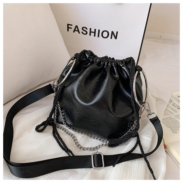 Black pleated shoulder bag with a faux leather texture, featuring a bucket shape, a chain strap, and a white label in the background with the text 'FASHION'.