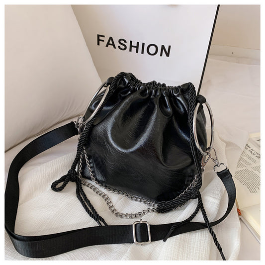 Black pleated shoulder bag with a faux leather texture, featuring a bucket shape, a chain strap, and a white label in the background with the text 'FASHION'.