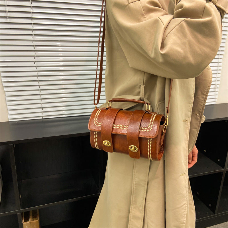 A person wearing a beige coat is holding a brown faux leather messenger bag with a white shoulder strap.
