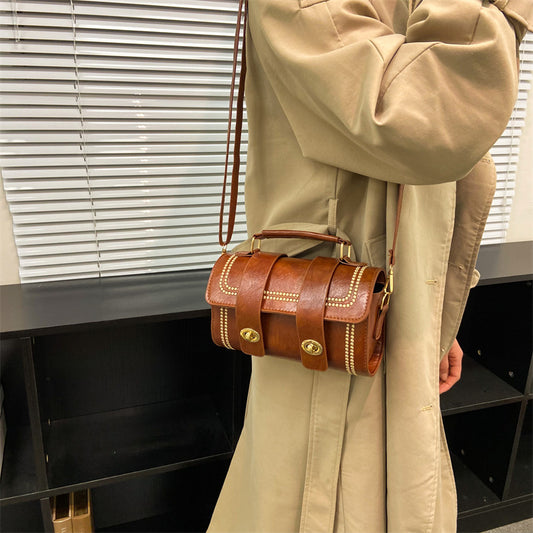 A person wearing a beige coat is holding a brown faux leather messenger bag with a white shoulder strap.
