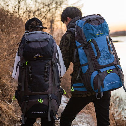 Two individuals hiking with large backpacks in what appears to be a cold environment, with trees and a body of water in the background.