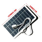 A portable solar panel charger connected to a black smartphone via a USB cable, with the solar panel laid flat on a dark surface.