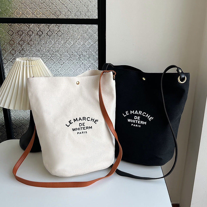 Two canvas shoulder messenger bags, one in white and one in black, with text printed on them, displayed on a table with a lamp and decorative items in the background.