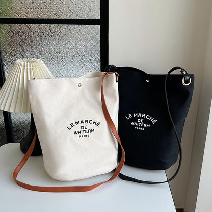 Two canvas shoulder messenger bags, one in white and one in black, with text printed on them, displayed on a table with a lamp and decorative items in the background.