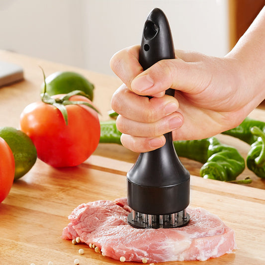 Hand holding a black meat tenderizer tool piercing a piece of meat on a wooden kitchen surface, with tomatoes and other vegetables in the background.
