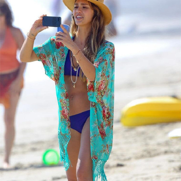 A woman at the beach wearing a green chiffon tassel coat with floral patterns and a bikini bottom, holding a mobile phone and taking a photo.