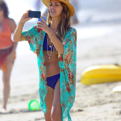 A woman at the beach wearing a green chiffon tassel coat with floral patterns and a bikini bottom, holding a mobile phone and taking a photo.