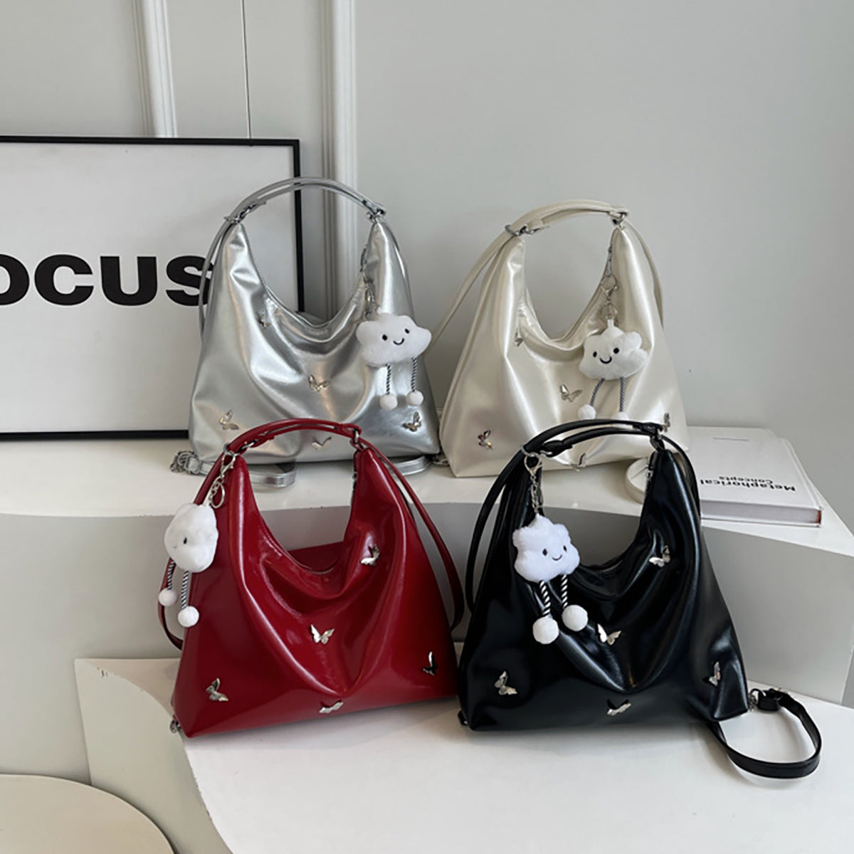 Four women's crossbody bags in silver, beige, red, and black colors, displayed on a table, with decorative tassels attached to them.