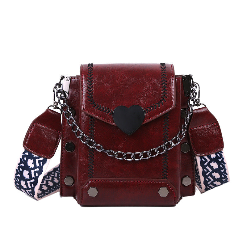 A red PU leather messenger bag with a chain shoulder strap, a heart-shaped clasp, and a blue patterned shoulder strap.
