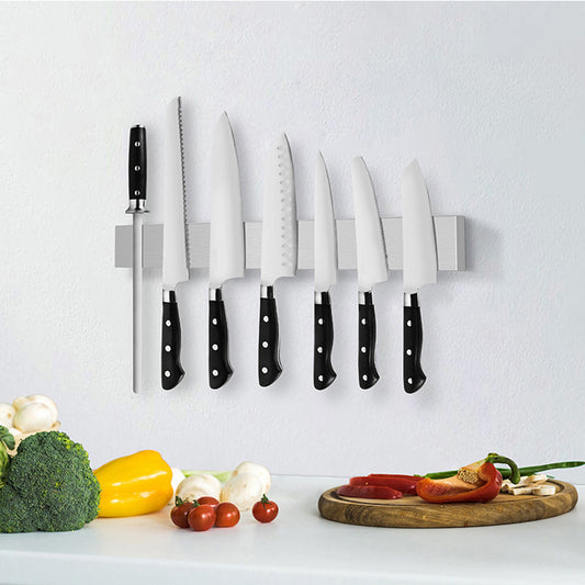 A set of various kitchen knives organized on a magnetic wall-mounted storage rack against a white background, with vegetables and a cutting board on the countertop in front.