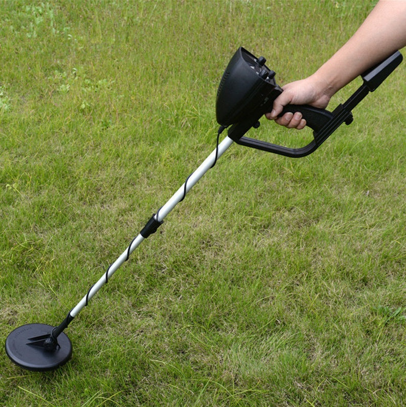 A hand holding a metal detector with a black handle and a circular probe, standing on grass.