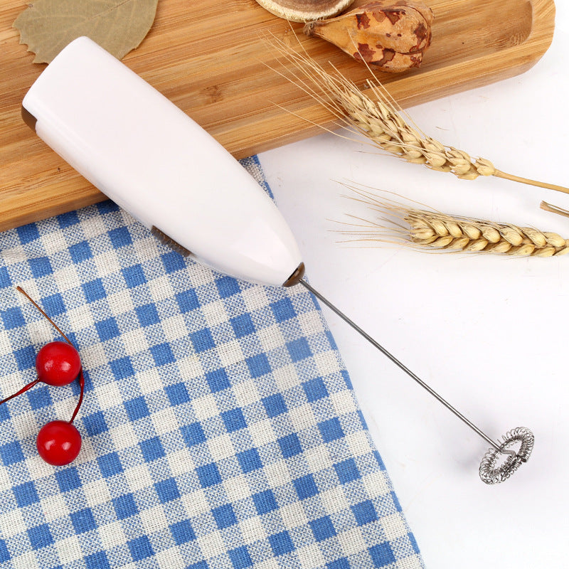 An electric coffee blender egg beater in white color, pictured on a table with a blue and white checkered napkin, some cherries, and wheat ears.