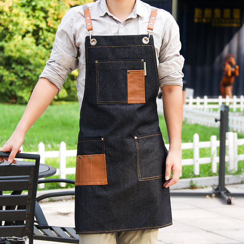 A man standing outdoors wearing a sleeveless black and blue apron with pockets and brown straps.