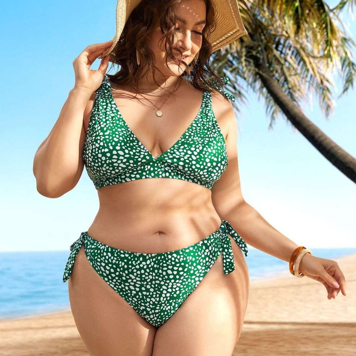A woman standing on a beach wearing a green bikini with a floral pattern, featuring a V-neck top and tie-side bottoms.