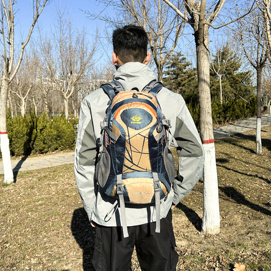 A person wearing a backpack with a solid pattern, featuring colors such as black, yellow, red, green, and khaki, in an outdoor setting with trees and a clear sky in the background.