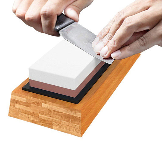 A person is sharpening a knife using a white corundum whetstone on a bamboo base.