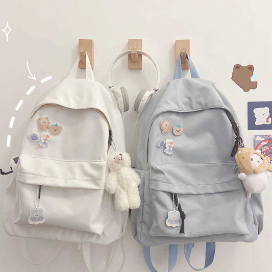Two backpacks, one in light blue and one in off-white color, adorned with various decorative items such as a plush toy and keychains, displayed against a wall with wooden hangers and a cartoon decoration.
