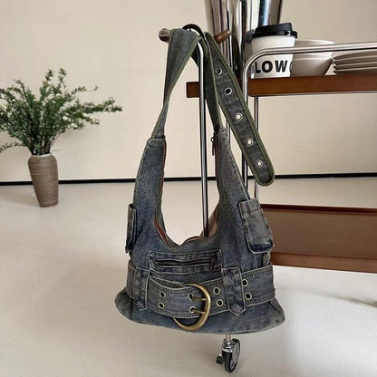 A denim shoulder bag with a single strap and metal hardware details displayed on a vertical stand, with a plant decoration in the background.